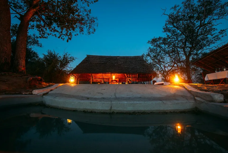 Thatched safari lodge lit by lanterns at dusk behind a stone-edged pool